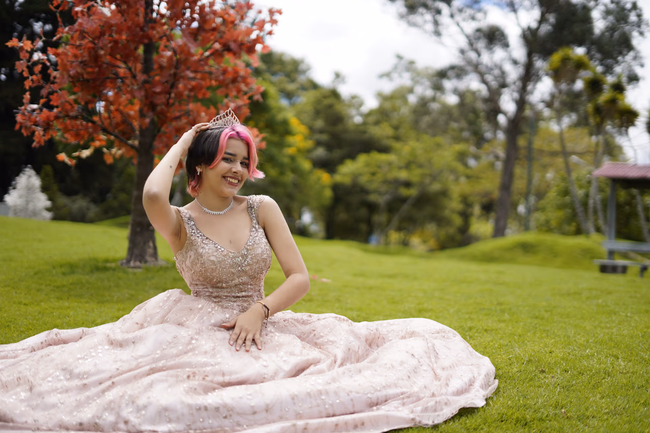 Joven con vestido rosado extendido sobre el césped, sonriendo mientras toca su tiara