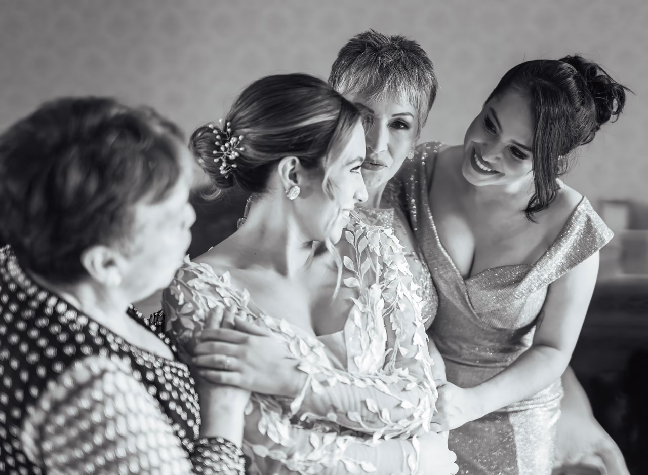 Novia sonriendo rodeada por tres mujeres durante sus preparativos, en blanco y negro
