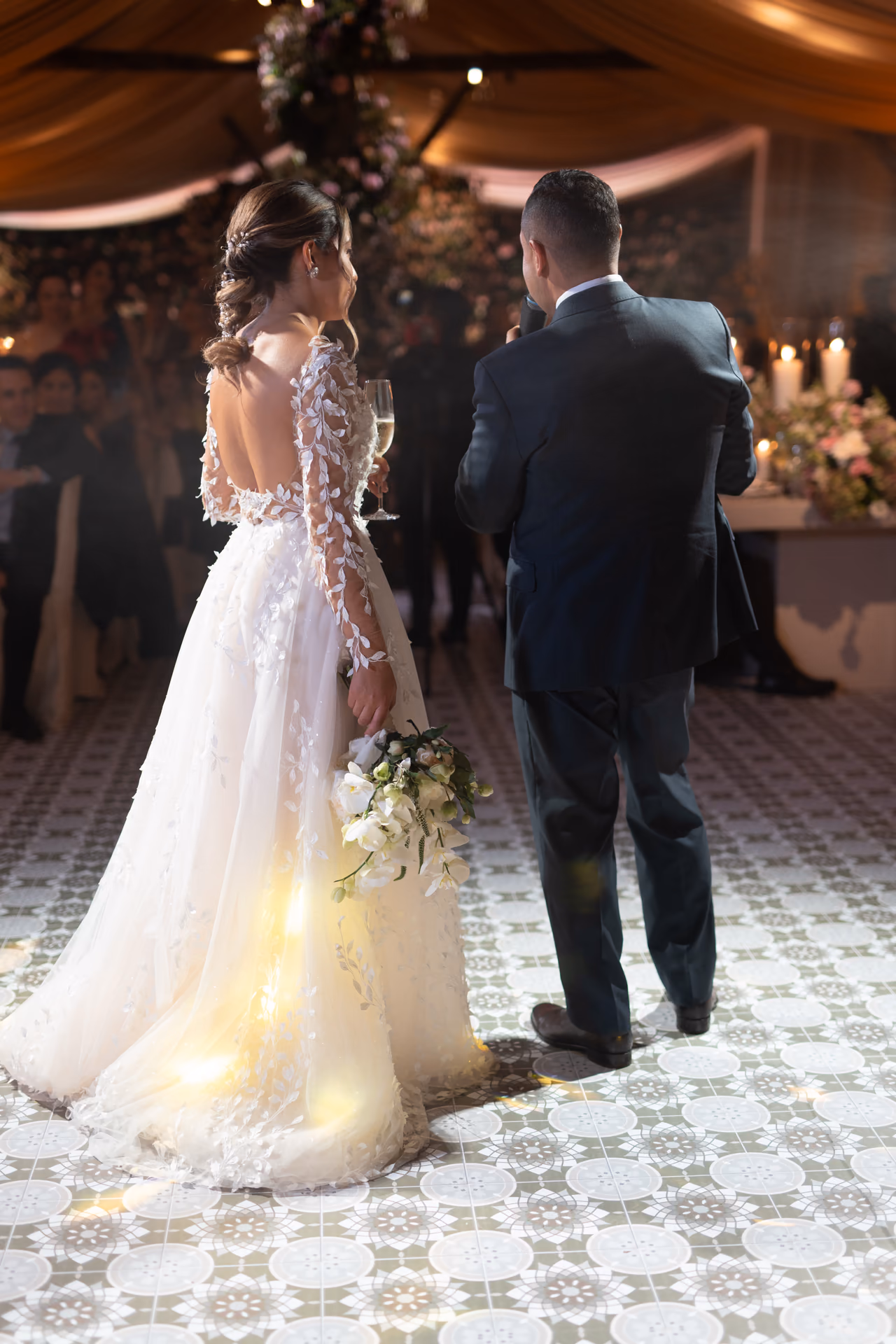 Novios de espaldas durante el brindis de su recepción, con luz sobre el vestido y el suelo
