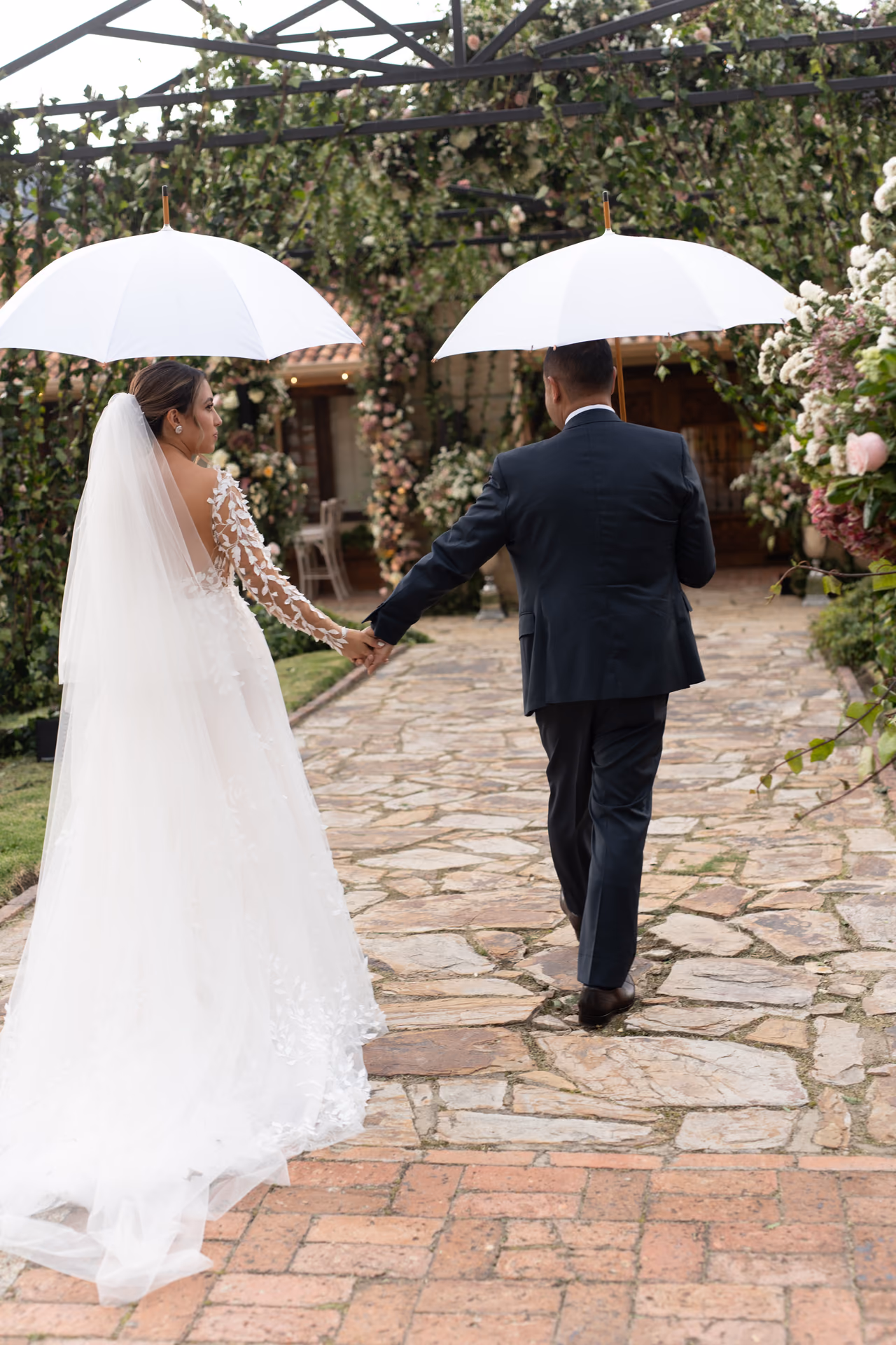 Novios caminando de la mano bajo dos paraguas blancos por un jardín con flores