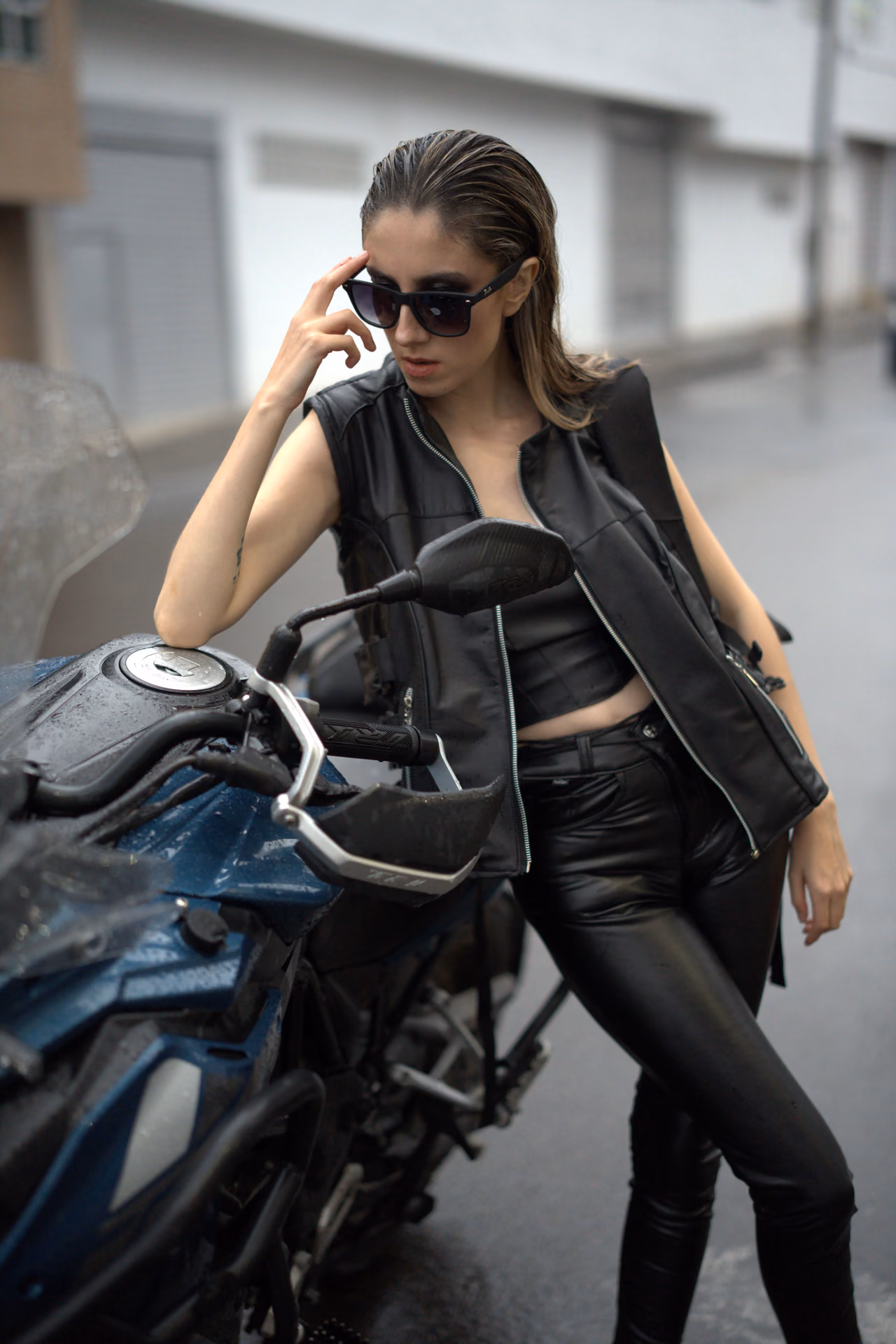 Modelo con chaleco negro ajustándose las gafas junto a una motocicleta azul en la ciudad