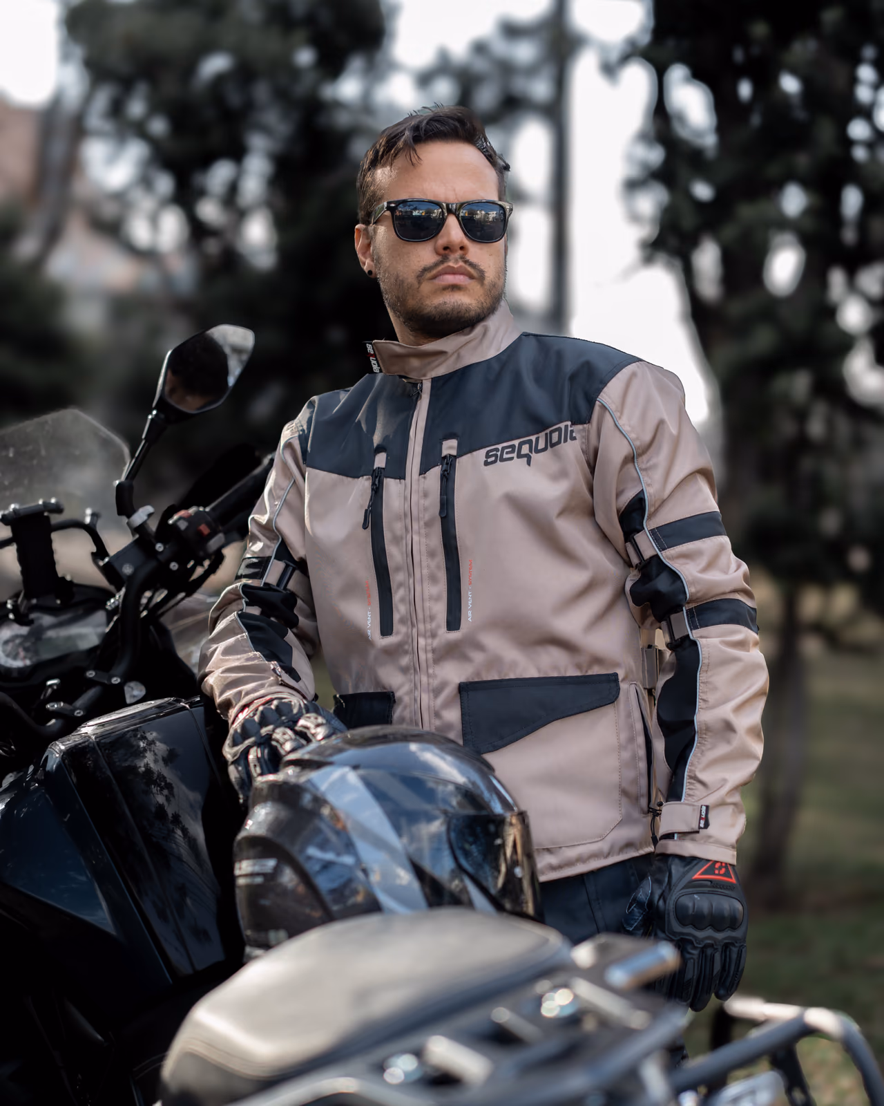Modelo con chaqueta protectora beige y gris, guantes y gafas junto a una motocicleta