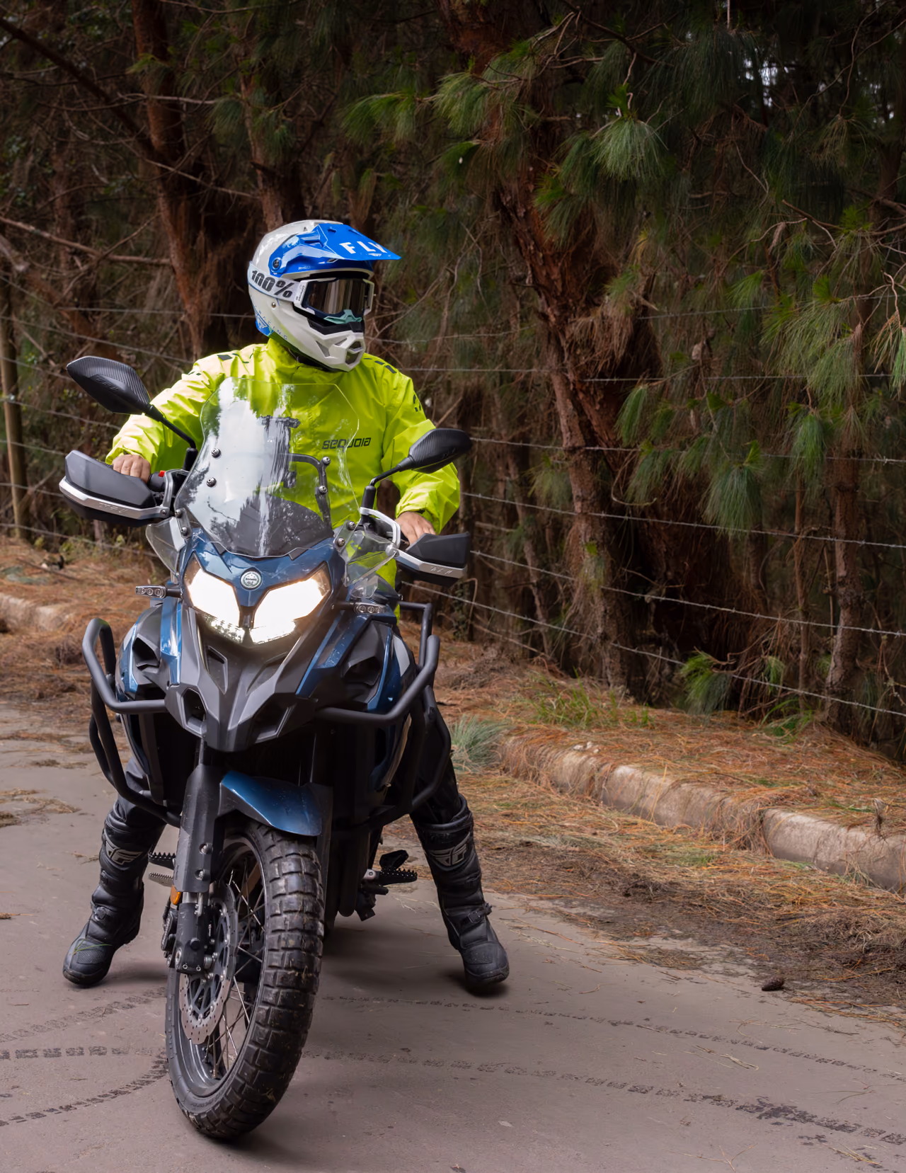 Motociclista con impermeable amarillo y casco azul sobre una moto junto a un bosque