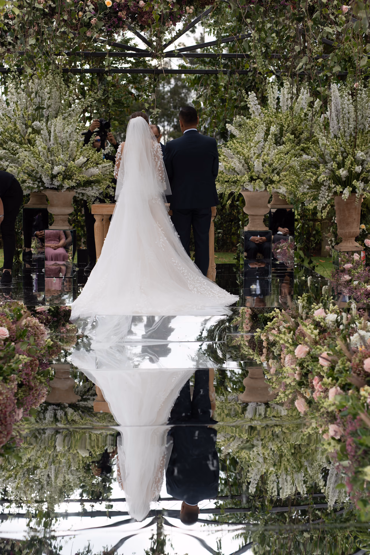 Novios de espaldas durante una ceremonia en jardín, reflejados en el pasillo de espejos